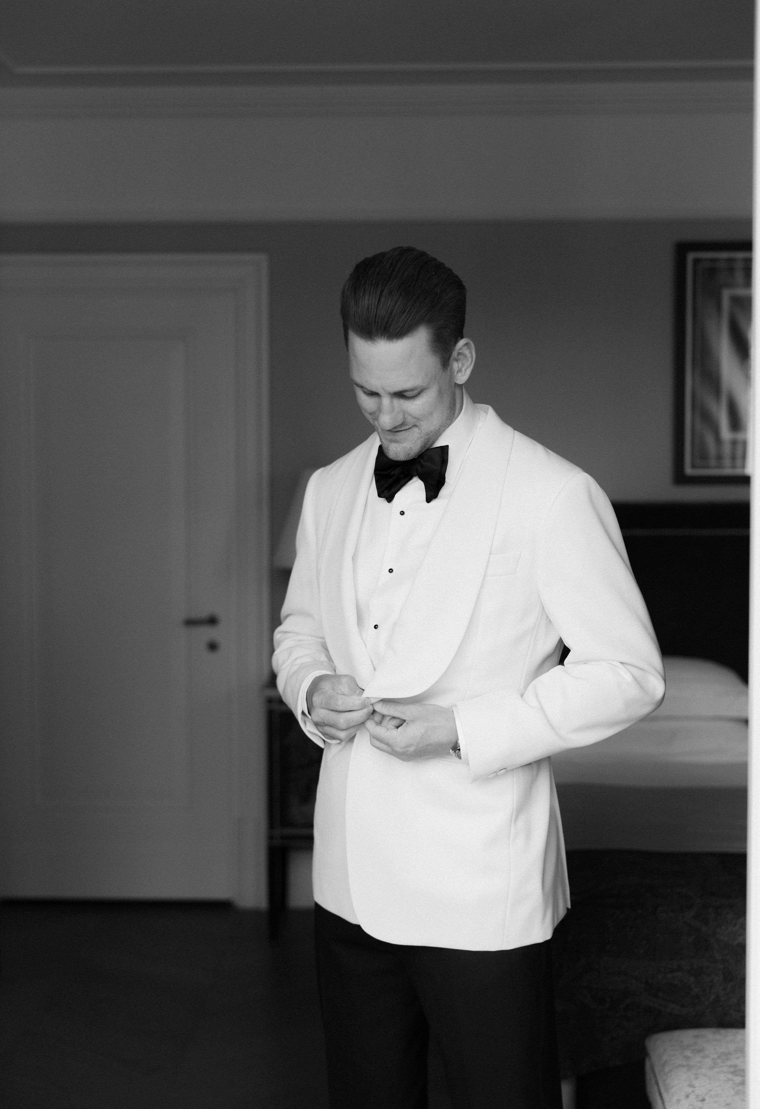 A person in a white tuxedo shirt and black bow tie adjusts their cufflinks in a black and white photograph.