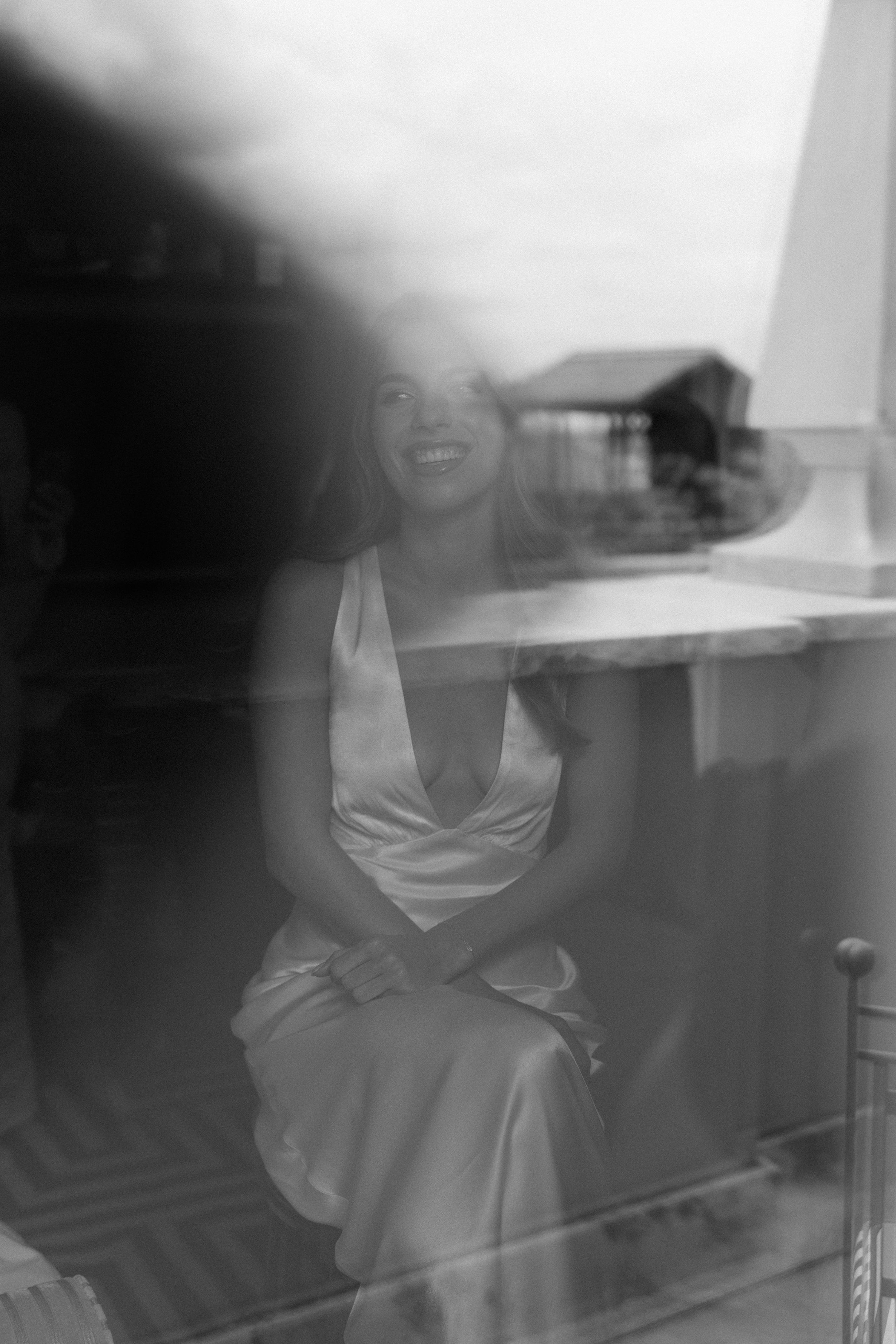 Dreamy black and white portrait through glass showing a person in a satin dress seated by a window.
