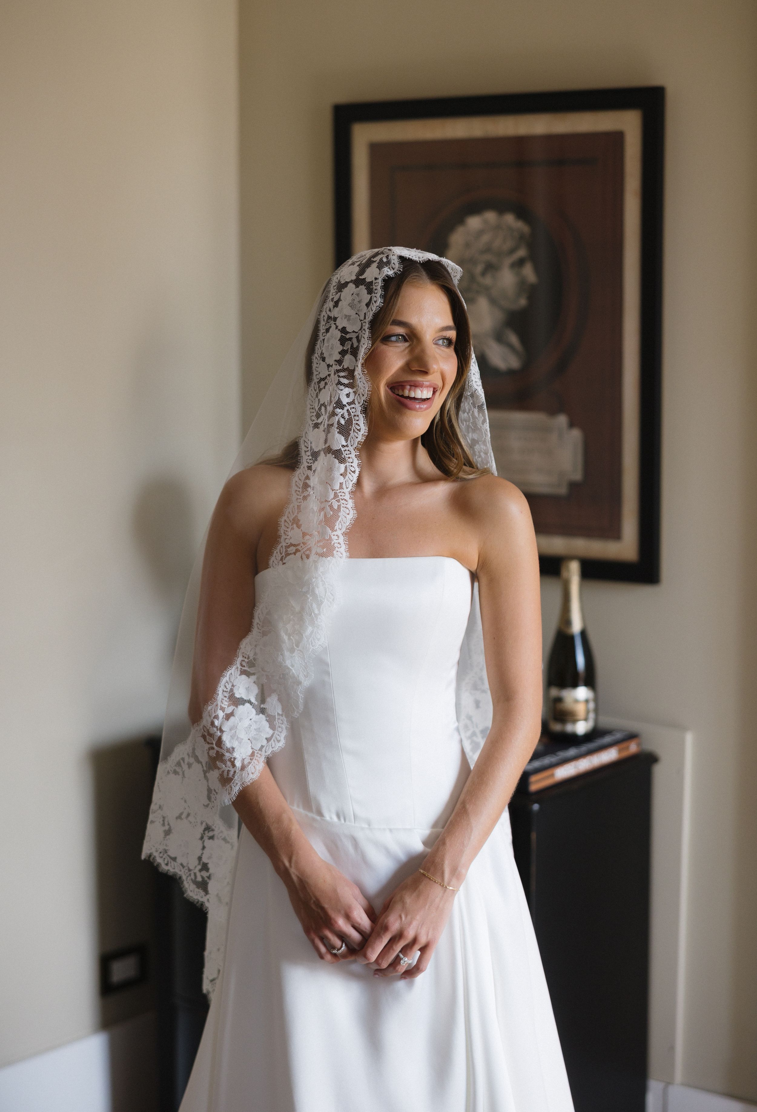 A bride in a classic white wedding dress and delicate lace veil stands elegantly in a softly lit room.