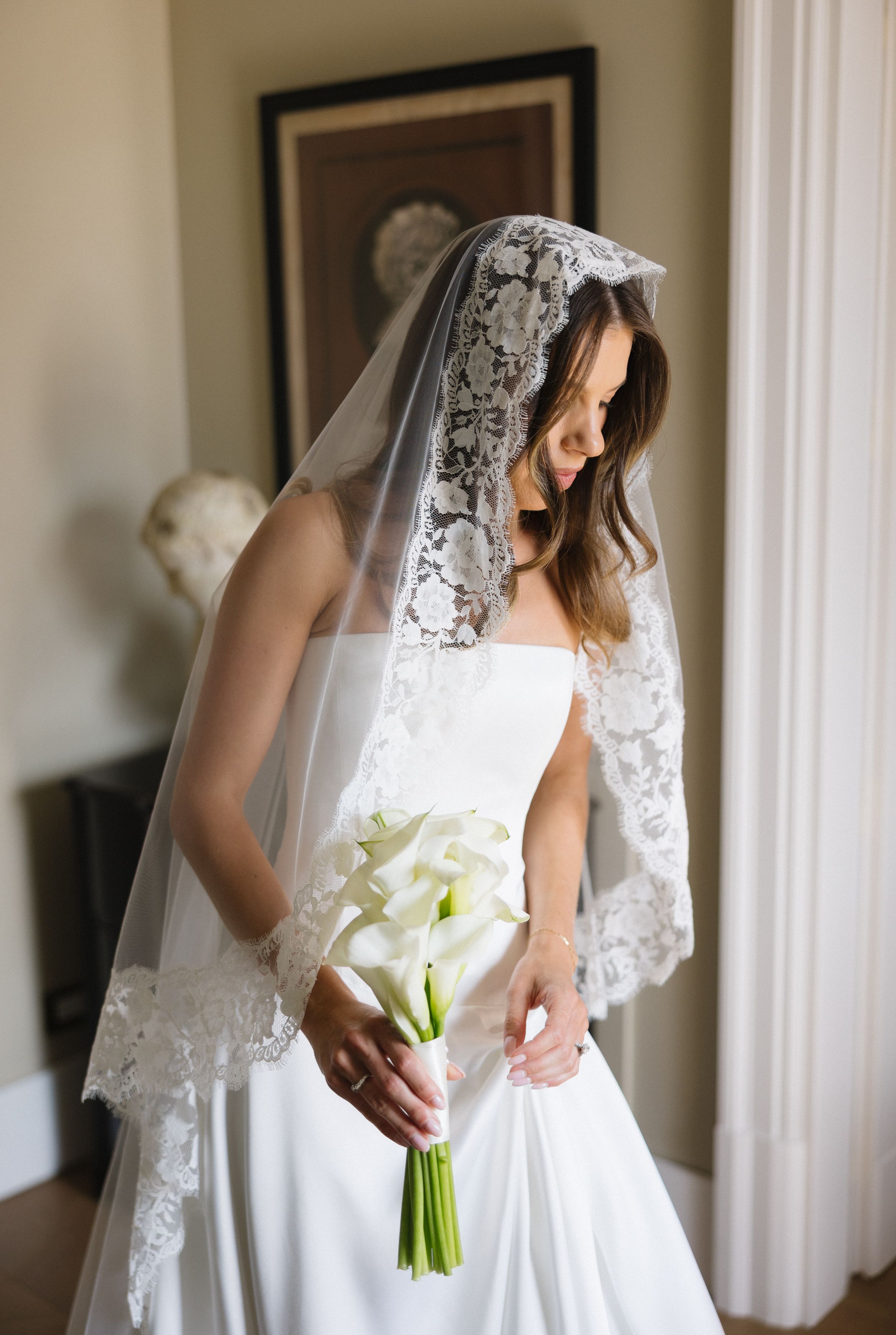 Bride in white wedding dress and lace veil holds a bouquet of white calla lilies near a window.