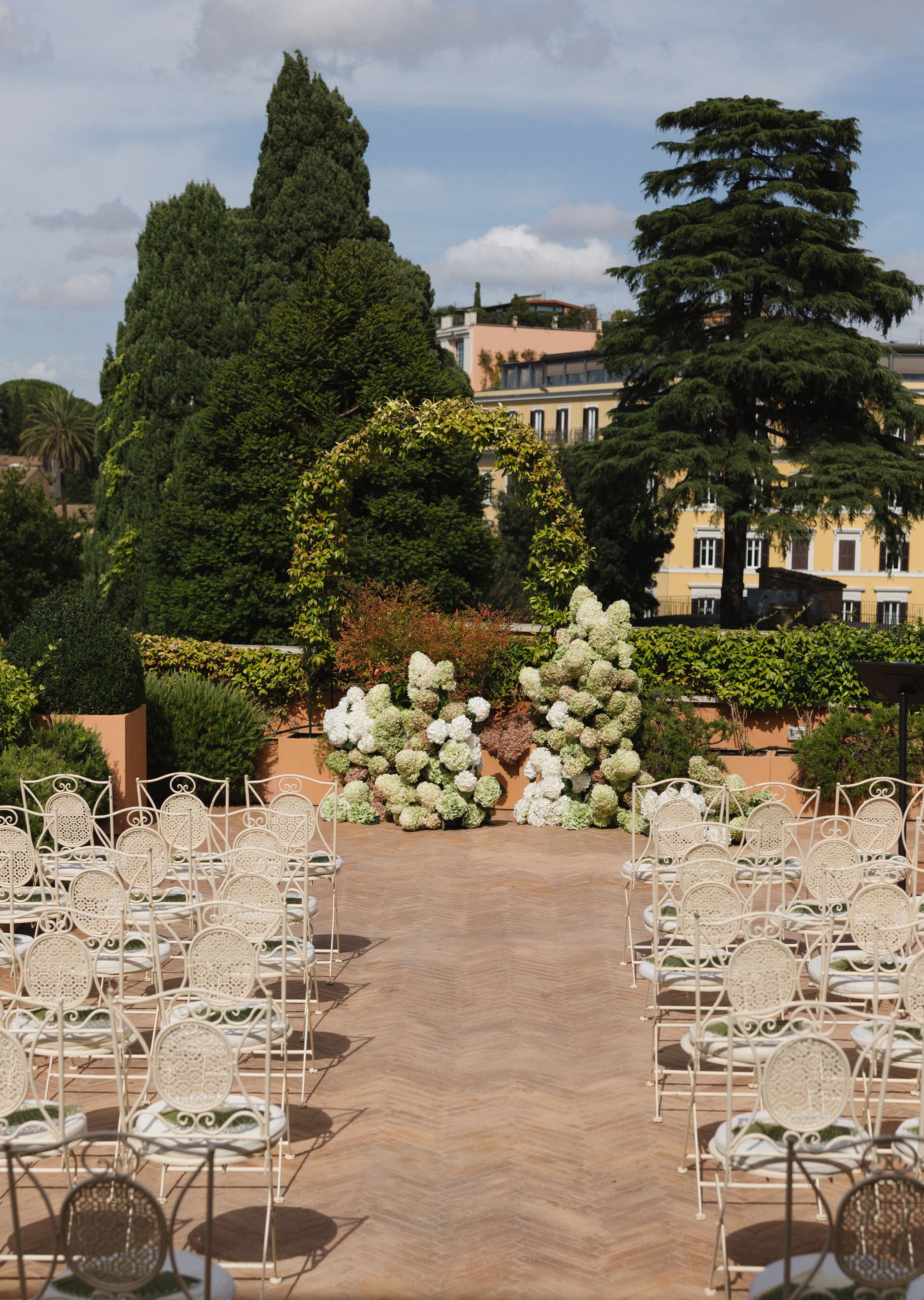 Elegant outdoor wedding venue setup with white chairs and floral arrangements on a terrace overlooking scenic gardens.