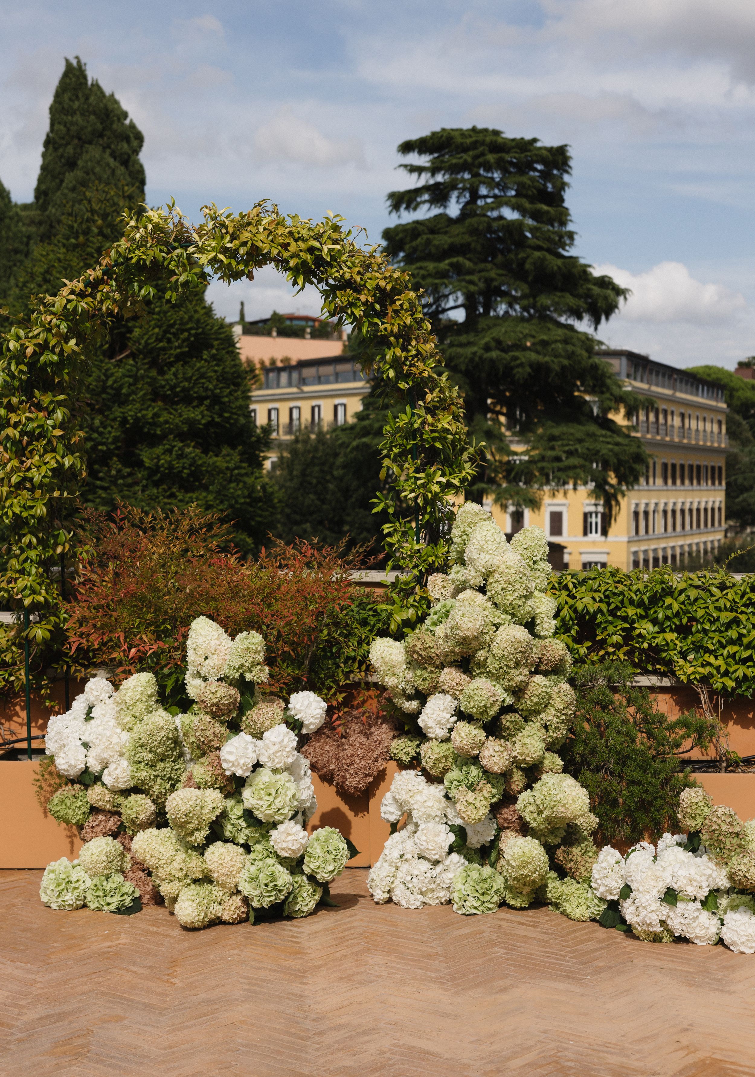 White hydrangea flowers arranged in a decorative border against a stone wall with trees and buildings in the background.
