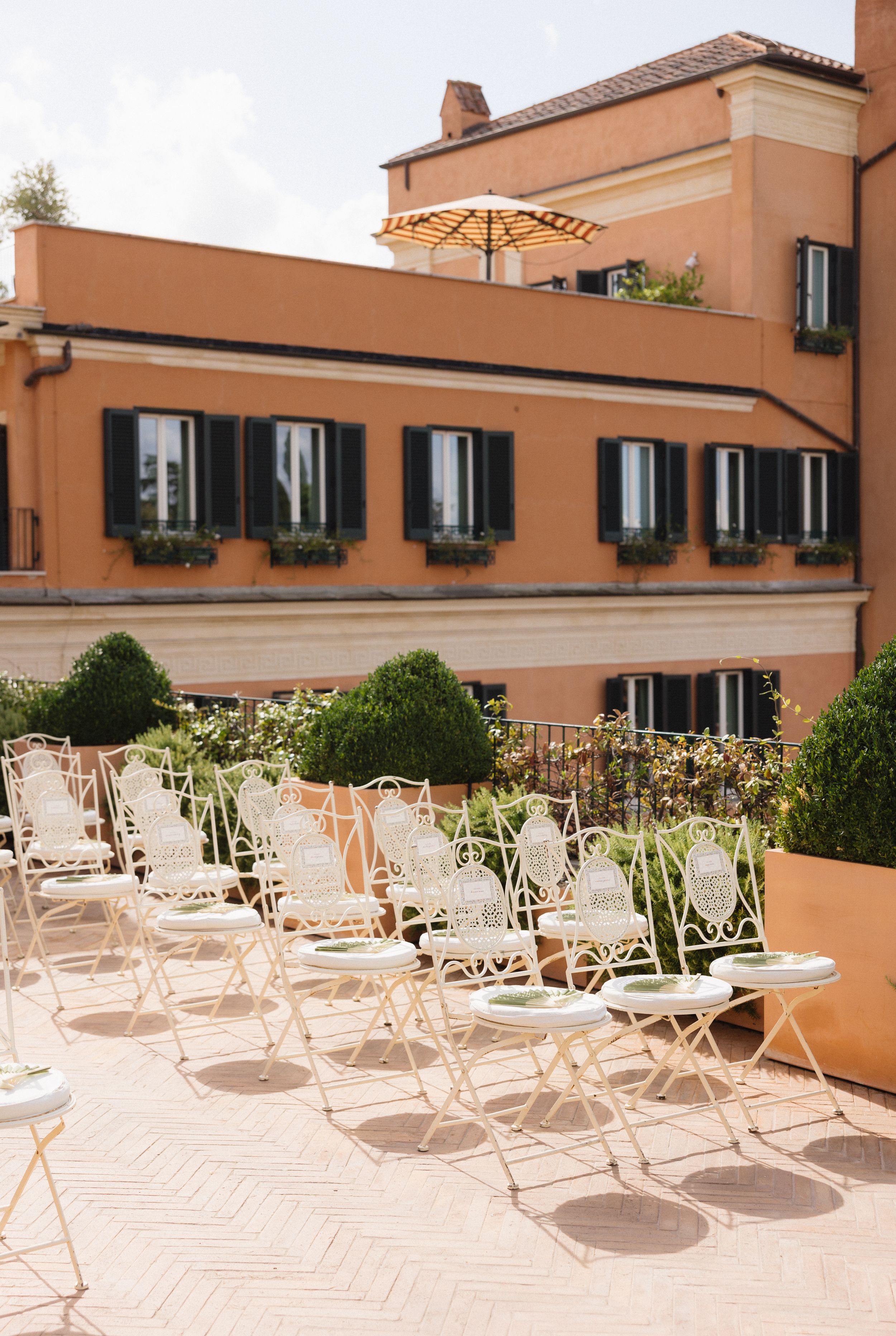 Empty white folding chairs arranged on a sunny terrace of a peach-colored Mediterranean building.