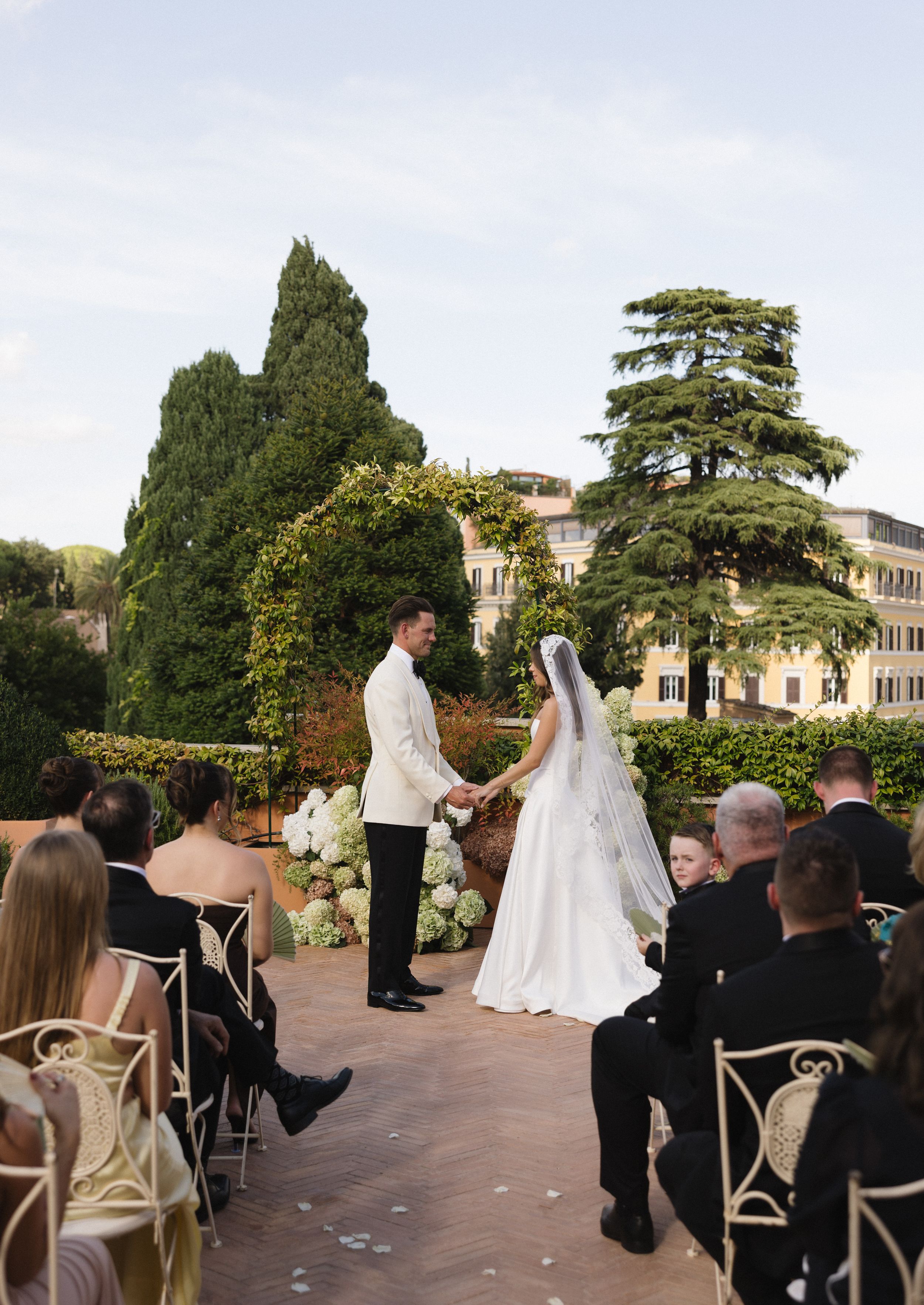 A wedding ceremony takes place on an elegant outdoor terrace surrounded by lush trees and architecture.