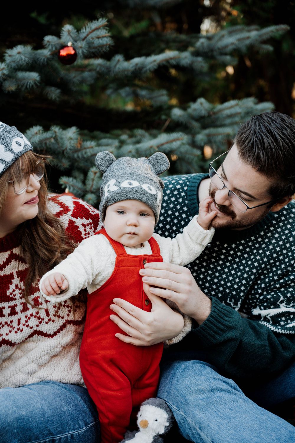 Winter family photoshoot in front of a decorated evergreen tree wearing matching knit sweaters and hats.
