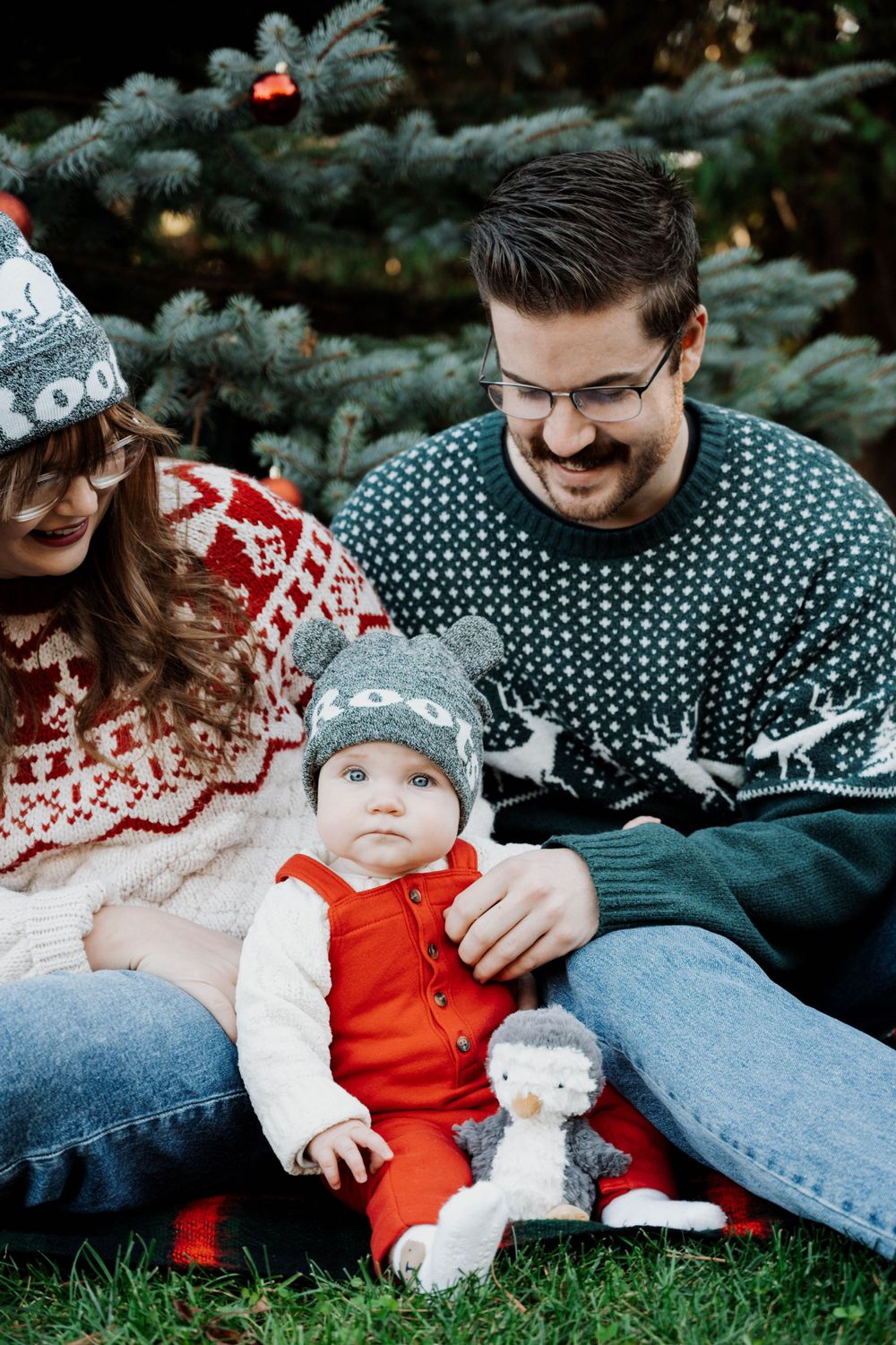 Family wearing cozy winter sweaters sits on grass by evergreen tree with small child in orange outfit and plush toy.