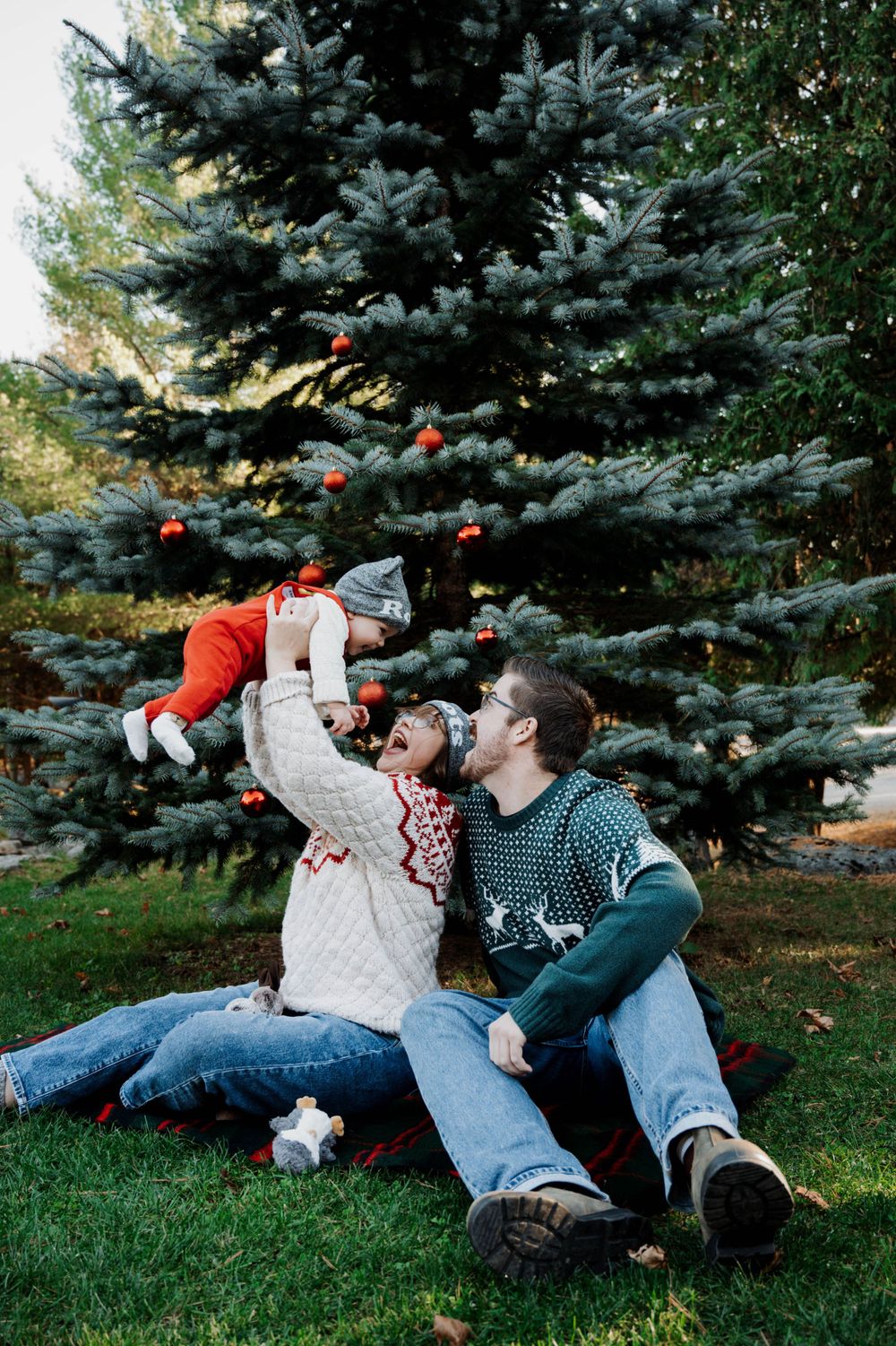 A couple sits together on grass beneath a large decorated blue spruce tree during a festive holiday setting.