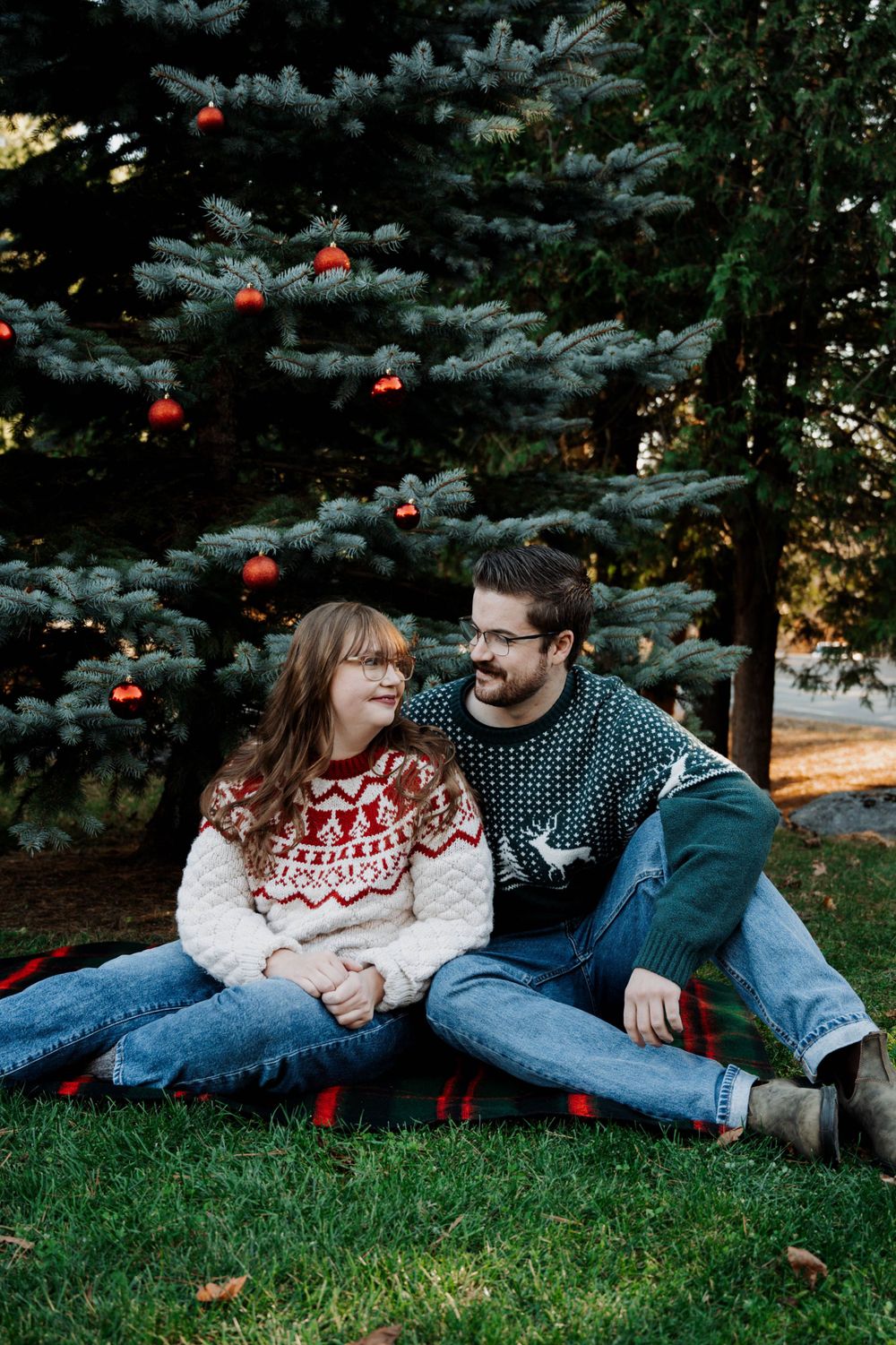 Two people in festive sweaters sit together by a decorated blue spruce tree with red ornaments.