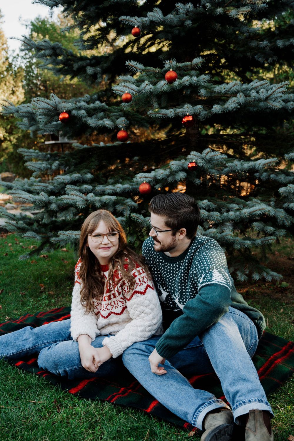 Couple in matching winter sweaters sitting on blanket beneath decorated blue spruce tree with red ornaments.