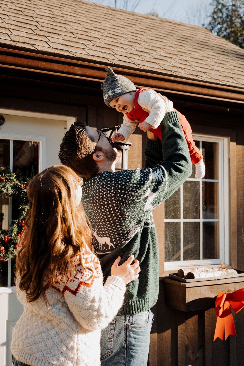 A family wearing cozy winter sweaters lifts their baby up high outside a rustic cabin during a bright sunny day.