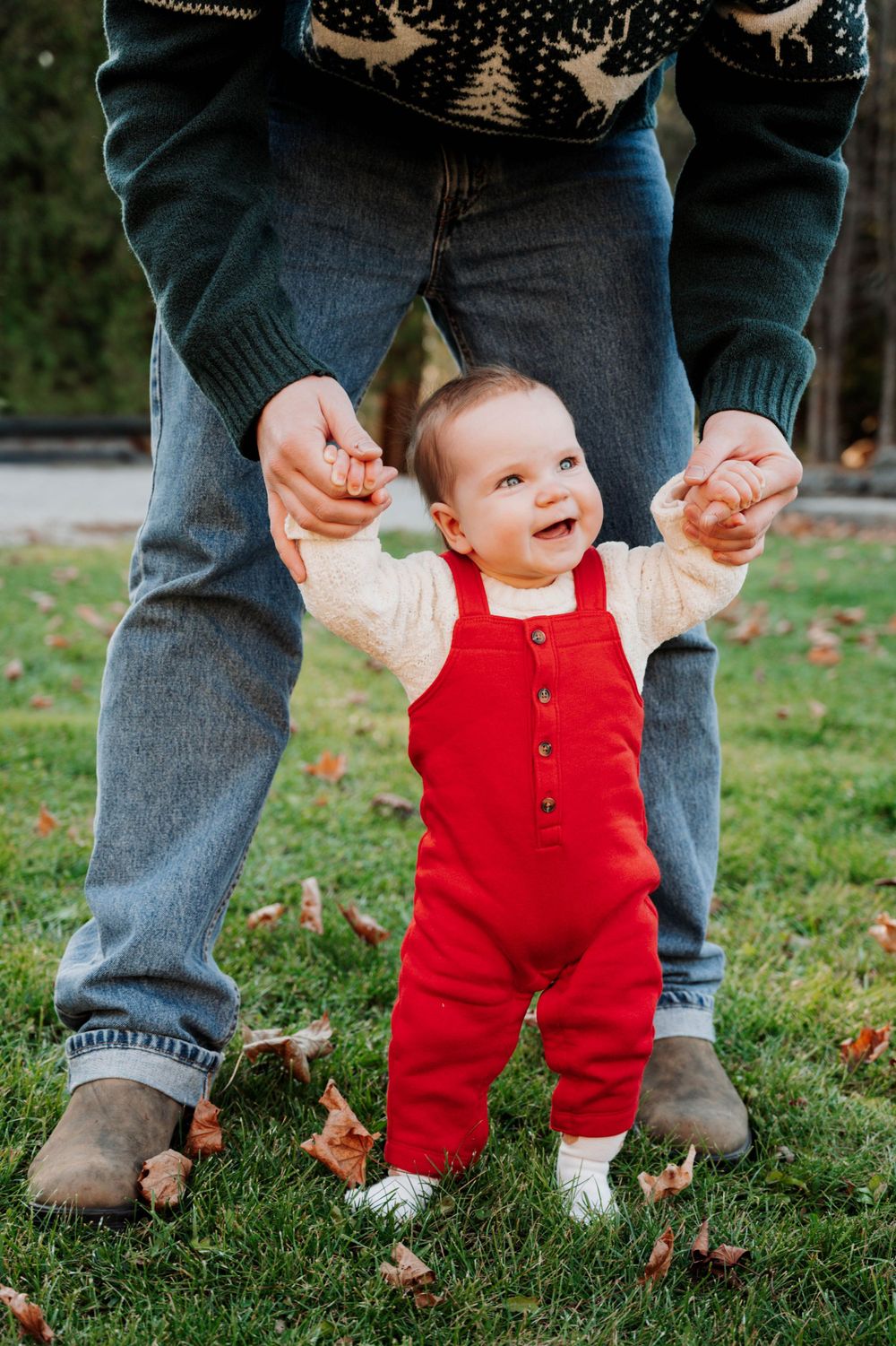Baby in bright red overalls stands on grass while holding onto adult wearing holiday sweater and jeans.
