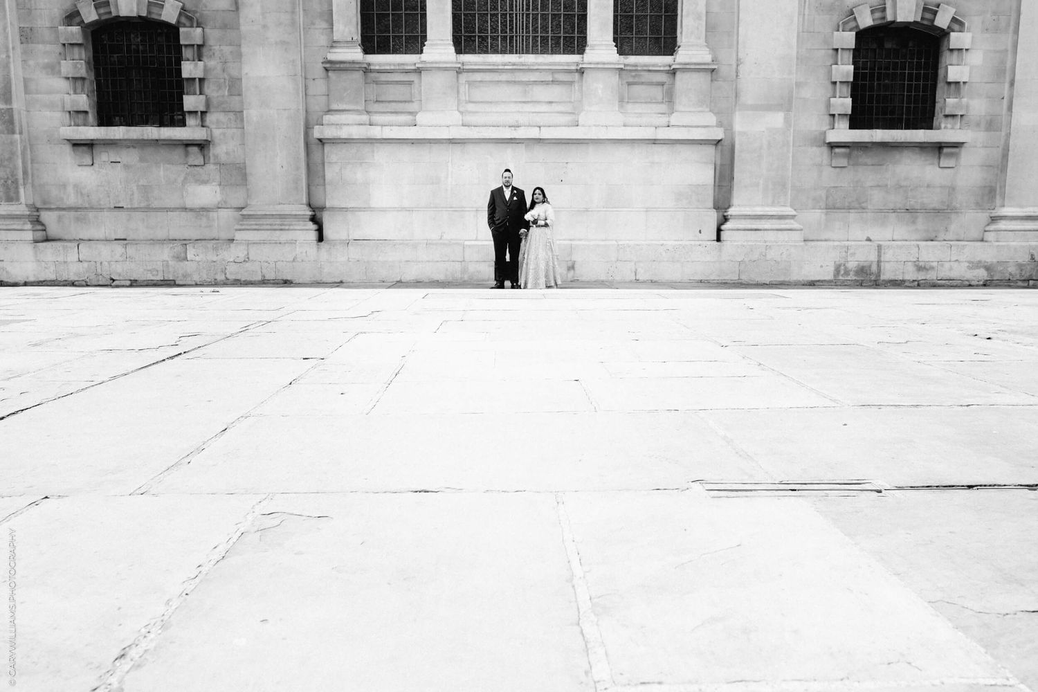 A black and white photograph of a couple standing against a grand white stone building with arched windows.