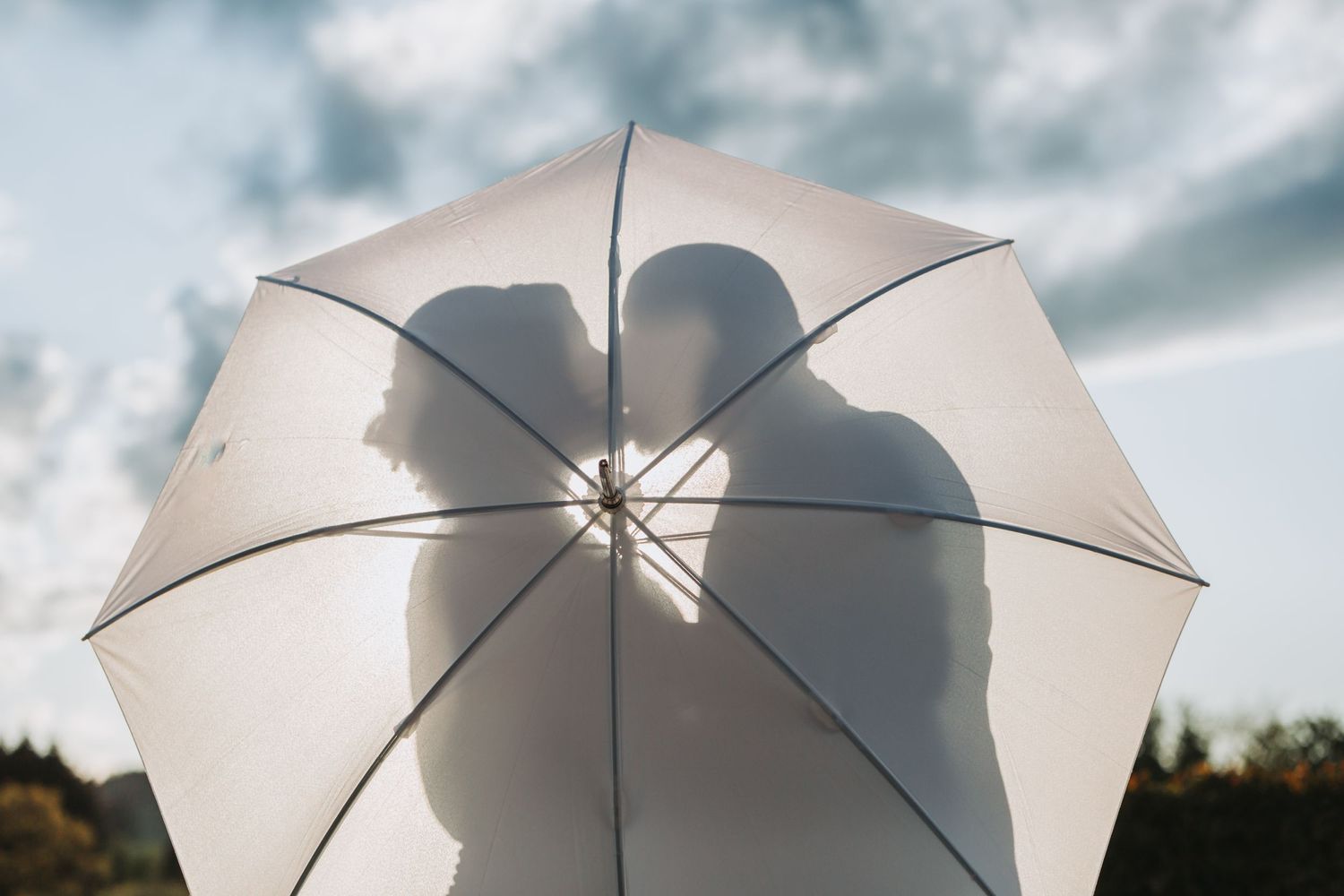 Silhouette of couple kissing behind a translucent white umbrella against a cloudy sky.