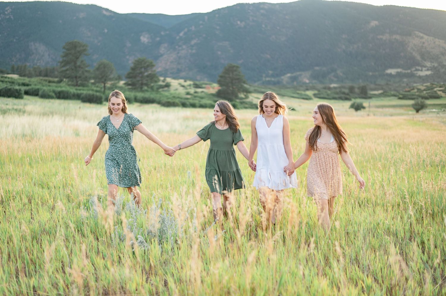 A mother and her adult daughters stroll through a summer field hand in hand in front of a mountain sunset
