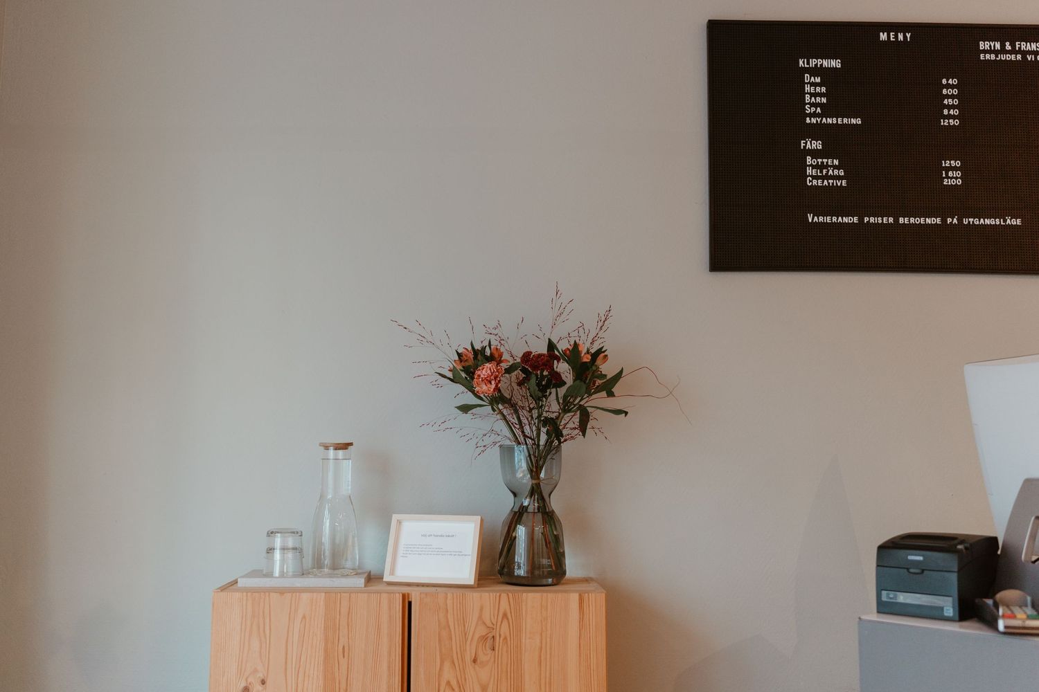 Dried floral arrangement in glass vase on wooden cabinet with minimalist decor and menu board mounted on wall.