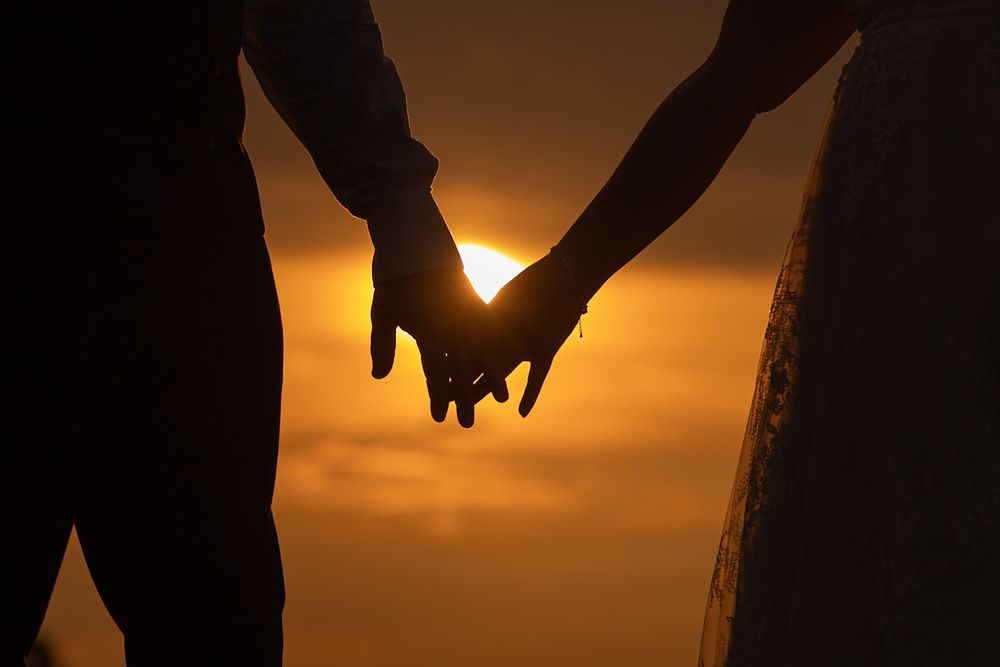 Silhouetted hands reaching towards each other against a dramatic orange sunset sky.