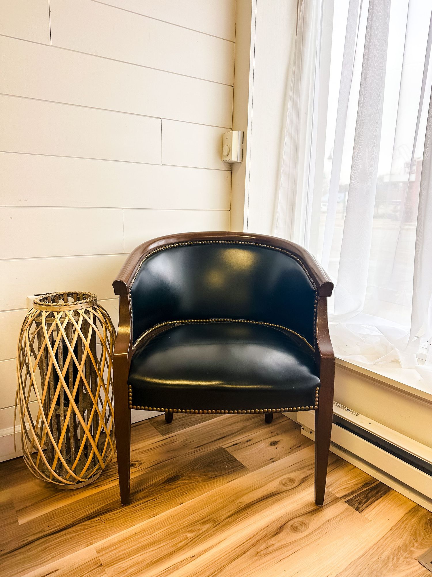 Vintage black leather barrel chair sits beside woven bamboo lantern on hardwood floor by bright window.