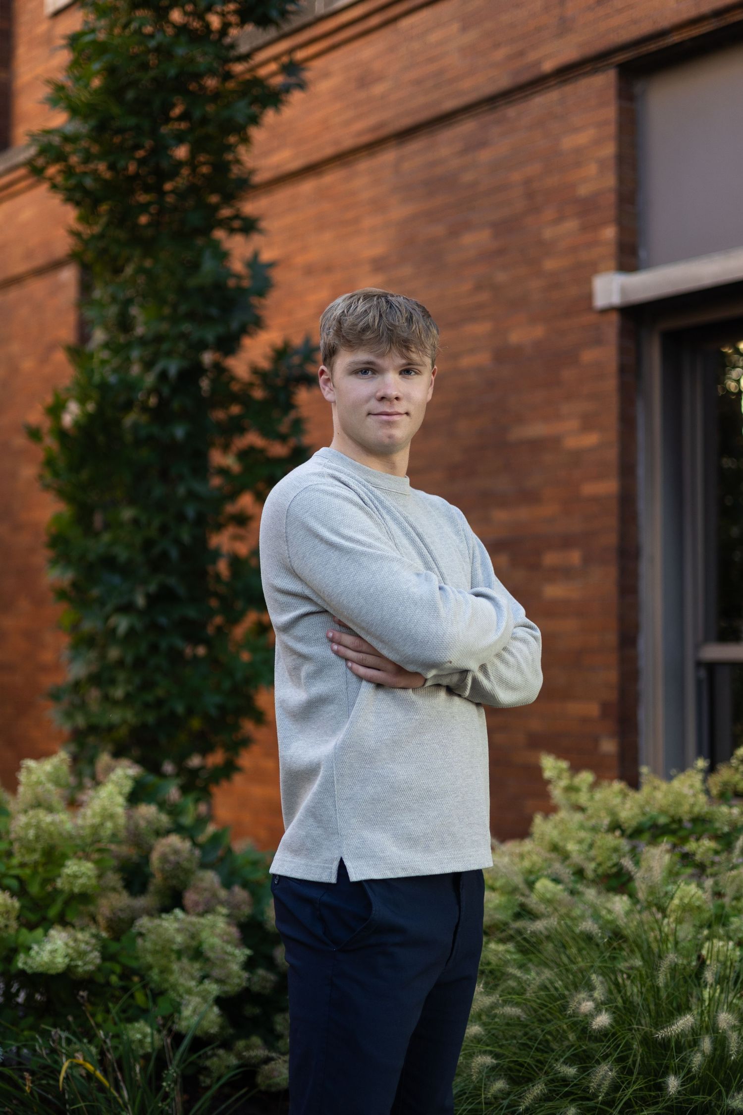 Portrait of someone wearing a light gray sweater standing in front of a brick wall with climbing vines and greenery.