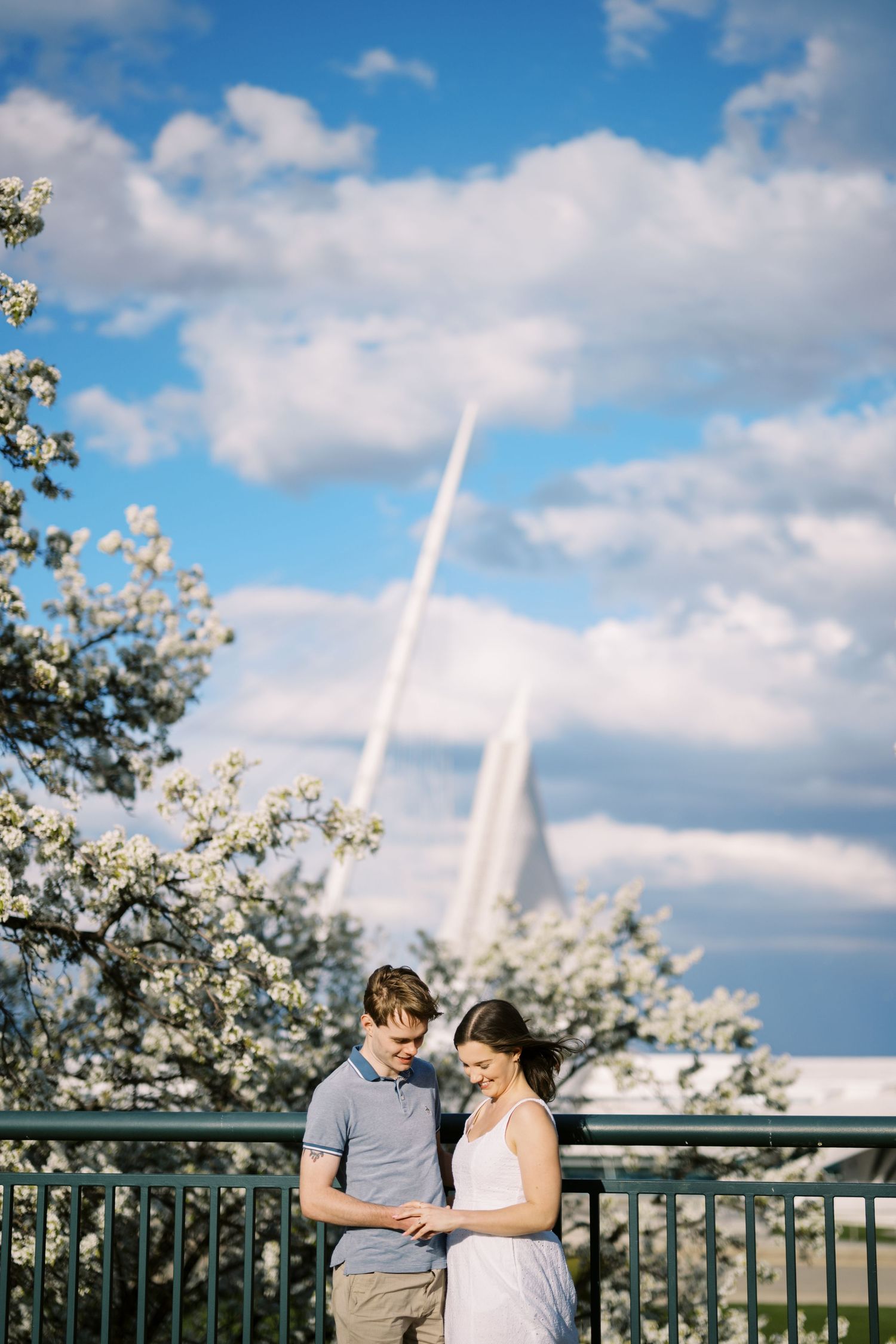 A romantic couple stands on a balcony with church spires and spring blossoms in the background on a sunny day.