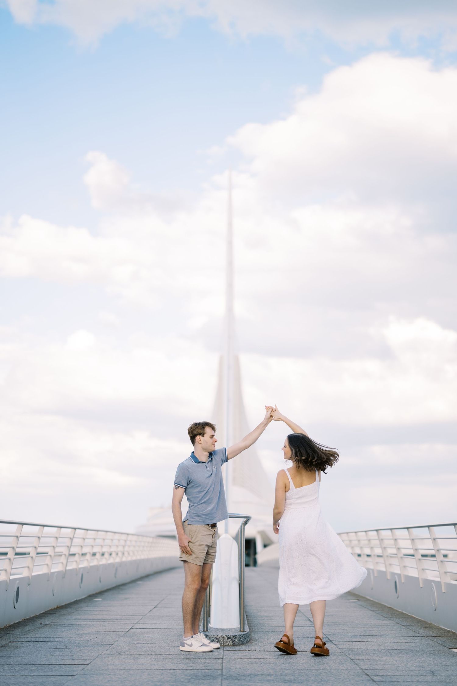 Couple dancing together on a modern white bridge walkway with a tall spire structure in the background.