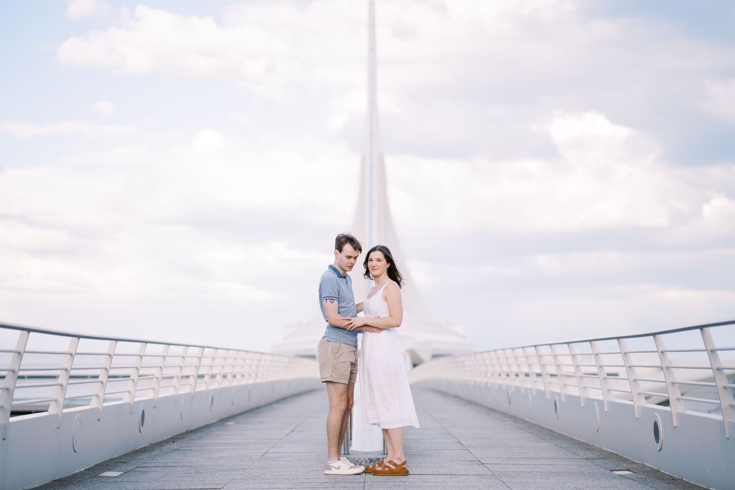 A couple stands together on a white modern bridge walkway against a bright cloudy sky.