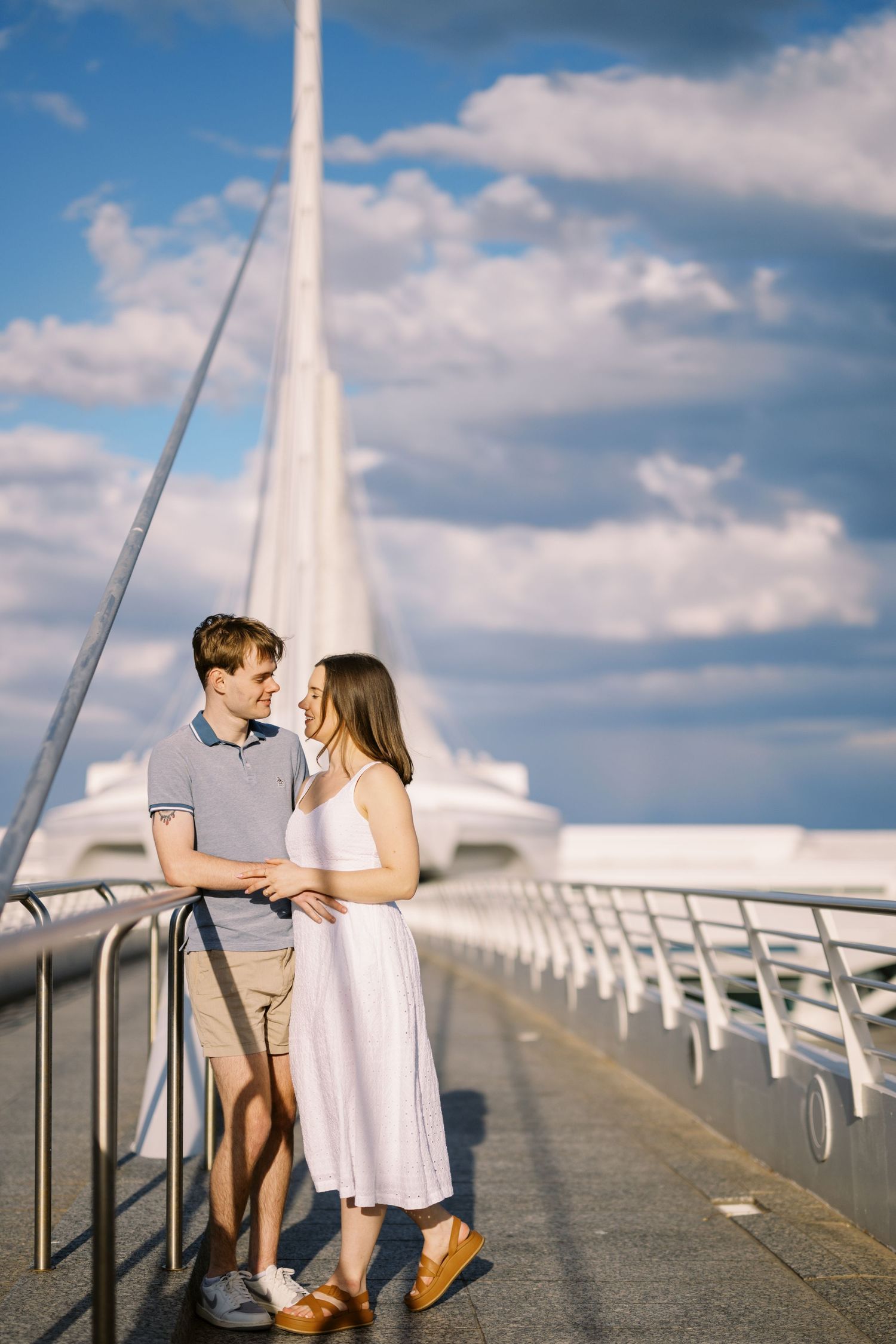 Romantic moment on a modern pedestrian bridge with striking architectural details and cloudy blue sky.