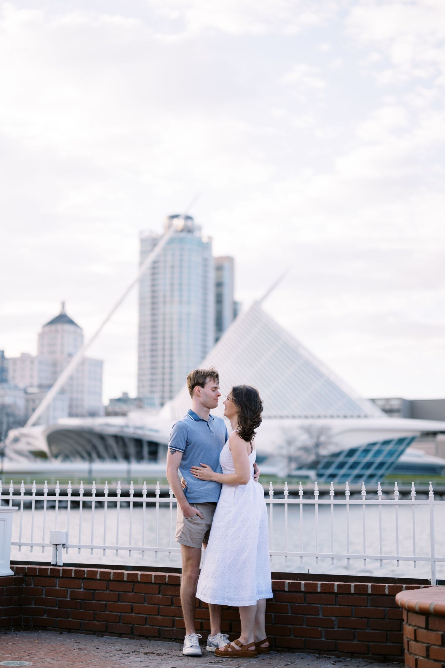 A couple stands on a brick walkway with Milwaukee's iconic Art Museum and downtown skyline visible in the background.