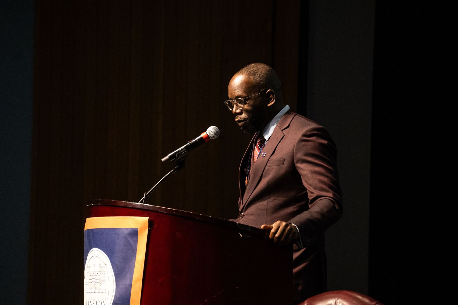 Speaker in a brown suit delivers remarks at a wooden podium with a microphone on a dark stage.