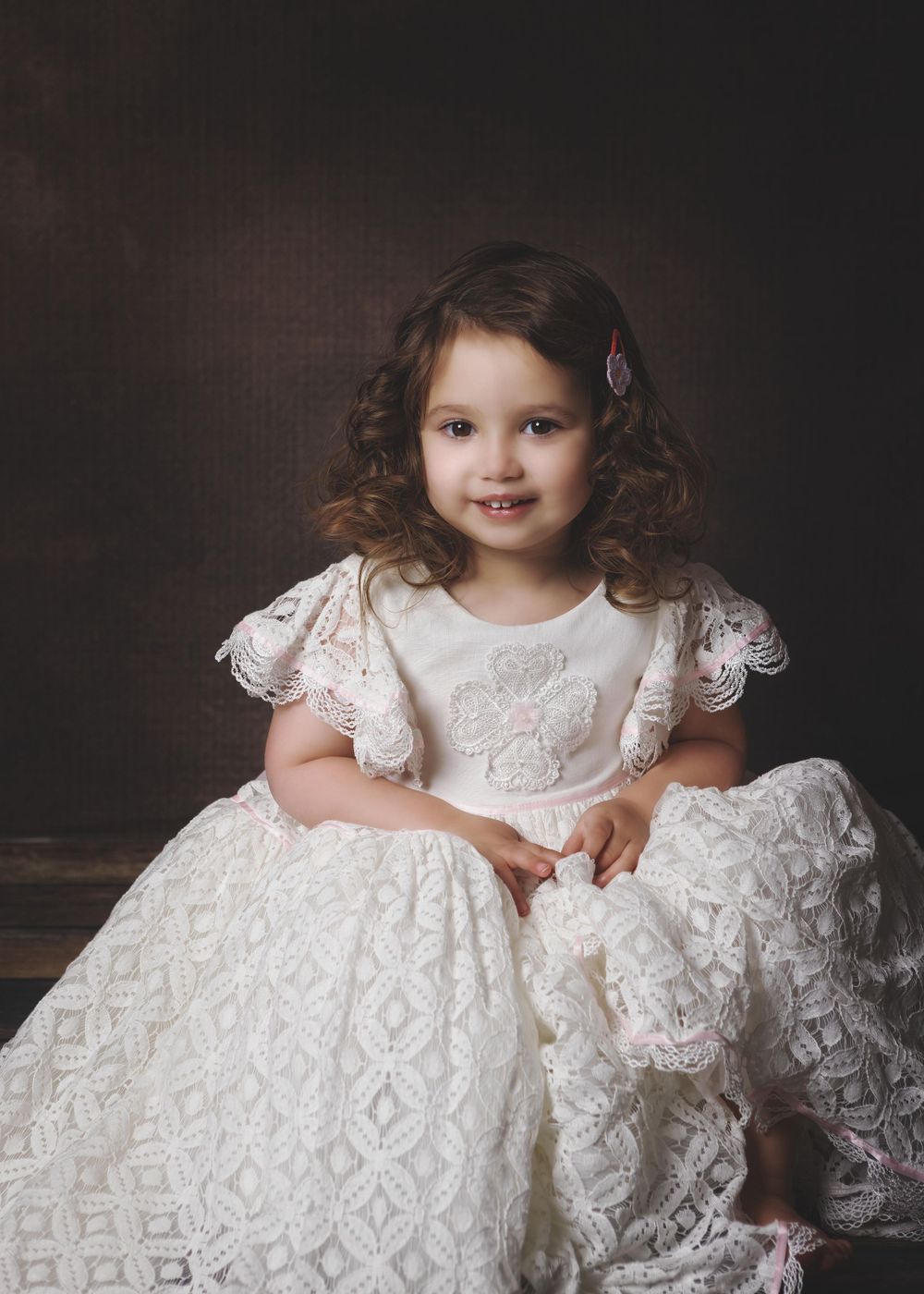 A child in a beautiful white lace dress with ruffled sleeves sits elegantly for a professional portrait photograph.