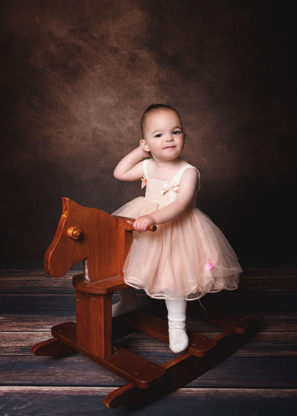 A baby in a pink tulle dress stands on a wooden rocking horse against a dark brown studio backdrop.