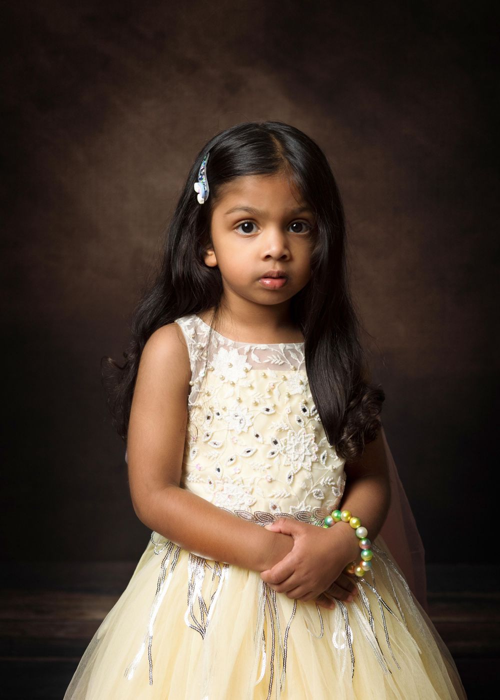 Portrait of young child in cream-colored tulle party dress with sequins against dark brown studio background.