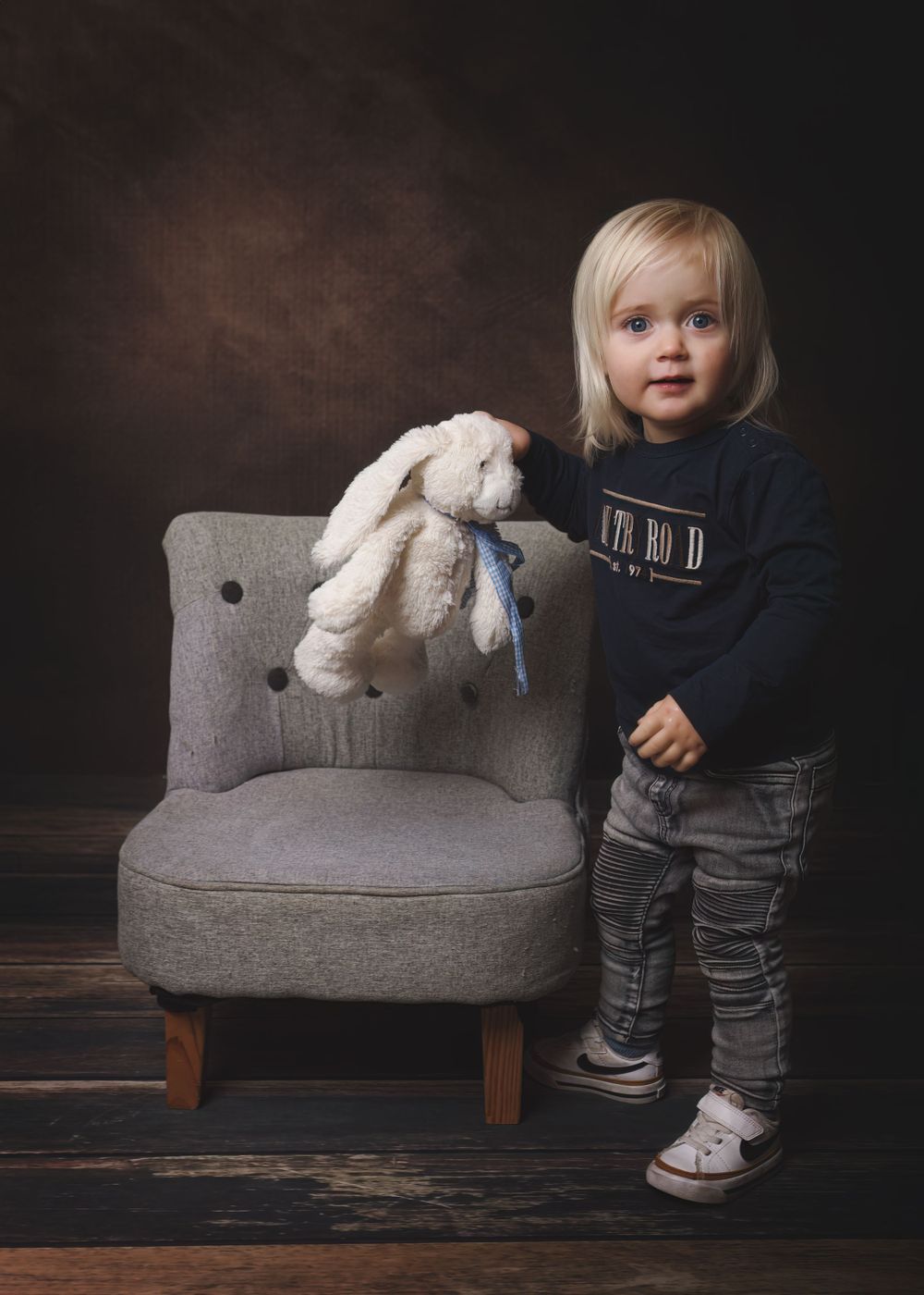 A small child stands next to a grey accent chair holding a plush white bunny toy in a dark studio setting.