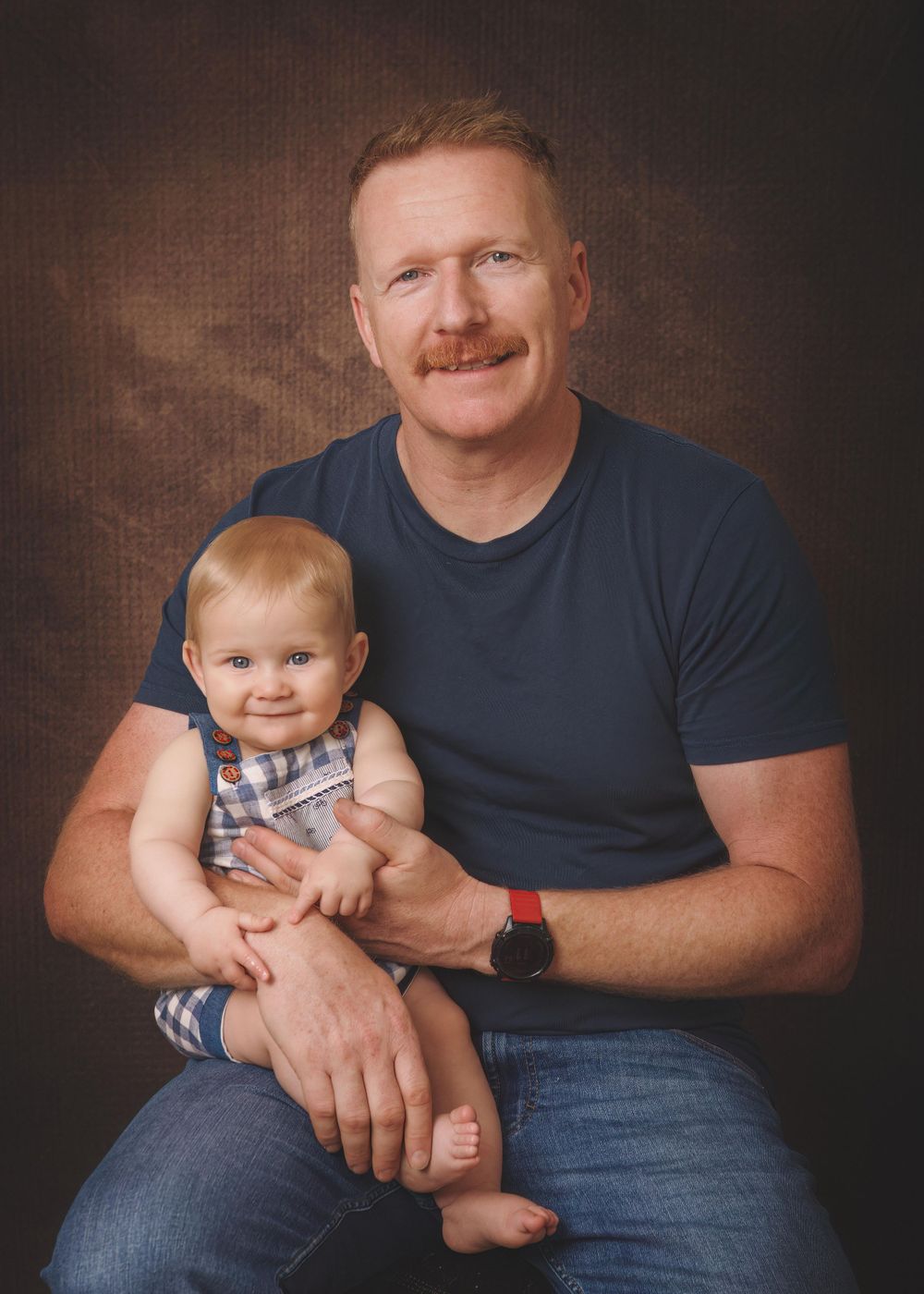 Portrait of an adult in a navy t-shirt holding a smiling baby wearing a plaid outfit against a brown backdrop.