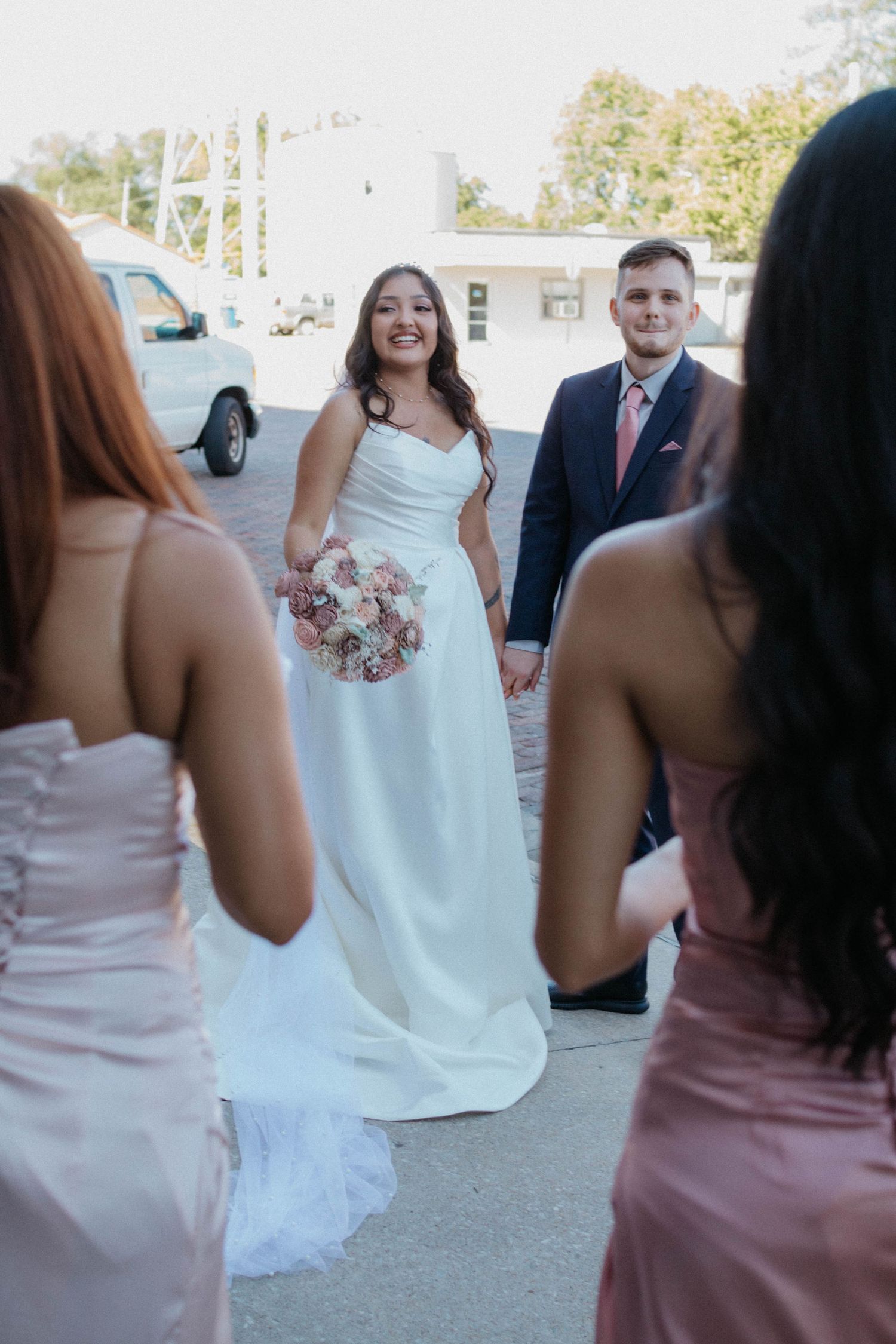 Wedding party celebrates outdoors with bridesmaids in pink dresses and couple in white dress and suit with bouquet. Candid wedding photo.