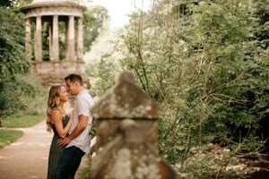A romantic couple shares an intimate moment on a walking path near a classical stone gazebo surrounded by lush greenery.