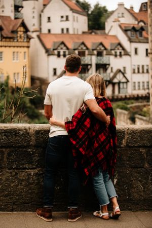 Two people lean on a stone wall overlooking charming European town buildings with timber-framed architecture.