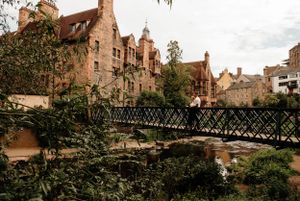 Historic stone buildings and iron footbridge over Dean Gardens in Edinburgh's scenic Dean Village on a cloudy day.