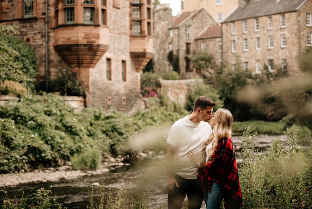 Romantic couple embraces in lush gardens beside historic stone buildings in Edinburgh's Dean Village area.