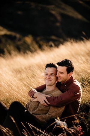 Two people share an intimate embrace while sitting in golden grass during sunset.