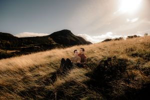 Two people sit in tall golden grass on a hillside at sunset with dramatic mountain silhouettes in the background.