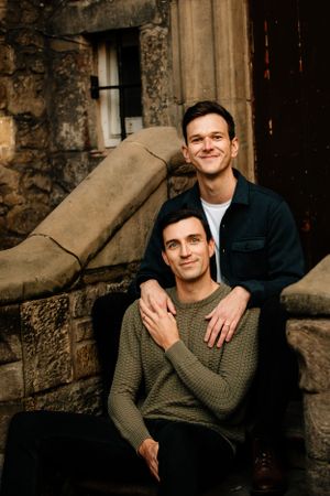A couple sits together on stone steps outside a rustic building with textured walls.