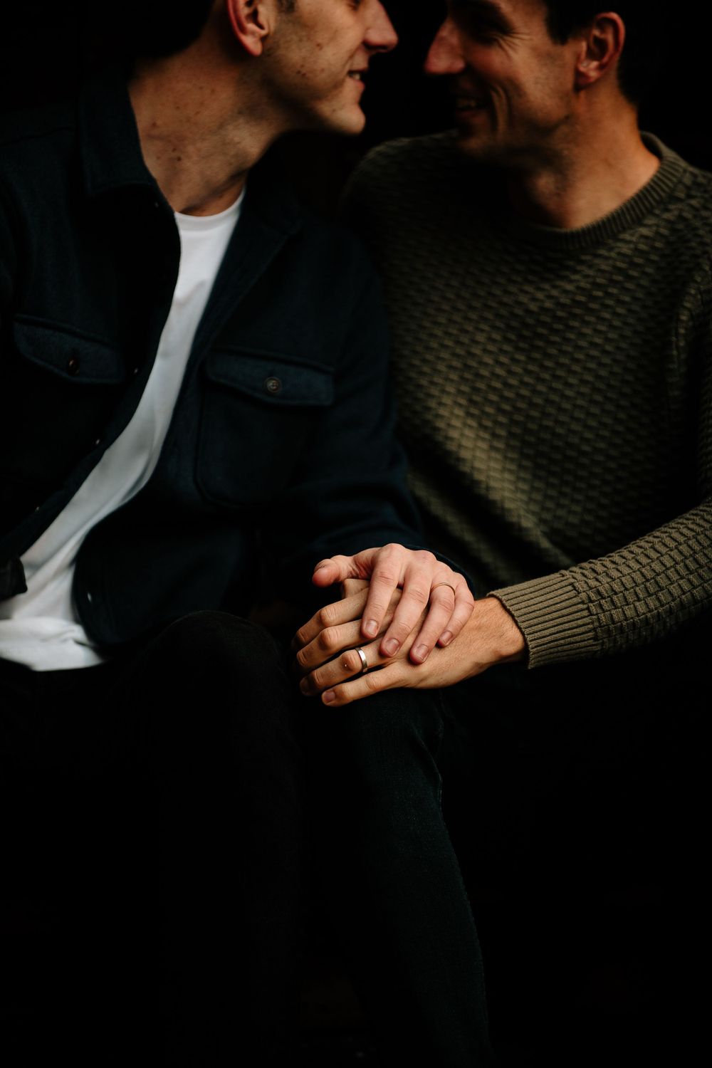 Two people in casual wear sitting close together holding hands intimately against dark background.