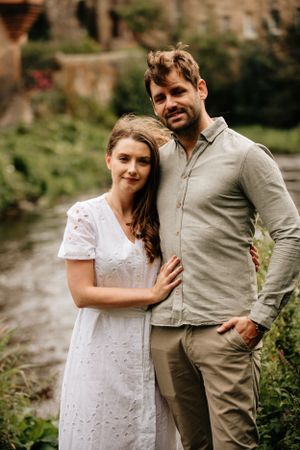 A couple stands together outdoors in casual attire, with her in a white lace dress and him in a gray button-down shirt.