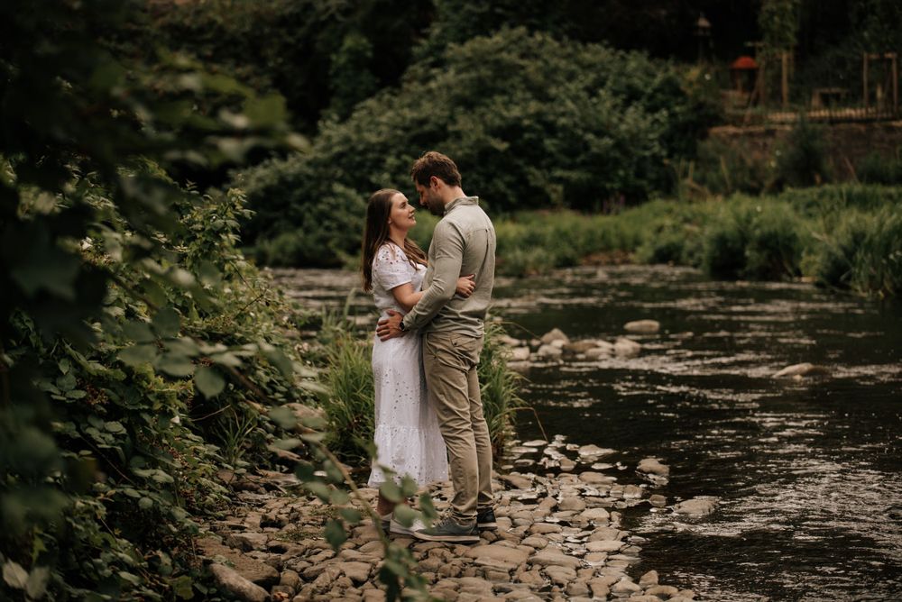 A romantic couple stands together on a rocky riverbank surrounded by lush greenery on a peaceful summer day.