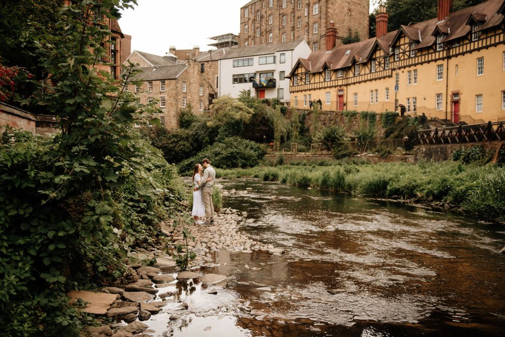 A figure in white stands on rocks by a stream with historic stone buildings and architecture in the background.