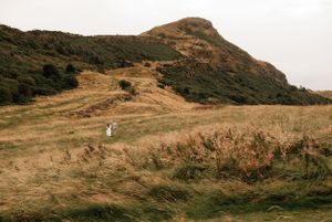 A winding dirt trail leads up a golden-brown hillside towards a rugged mountain peak under cloudy skies.