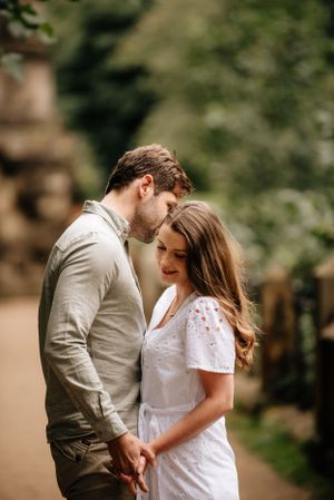 A romantic couple shares an intimate moment outside on a path surrounded by soft greenery and natural lighting.