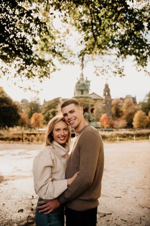 A couple embraces outdoors on an autumn day with a historic building and fall foliage visible in the background.