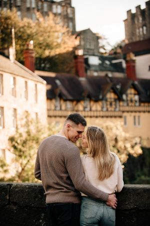 A couple shares an intimate moment against the backdrop of historic Edinburgh architecture in warm sunlight.