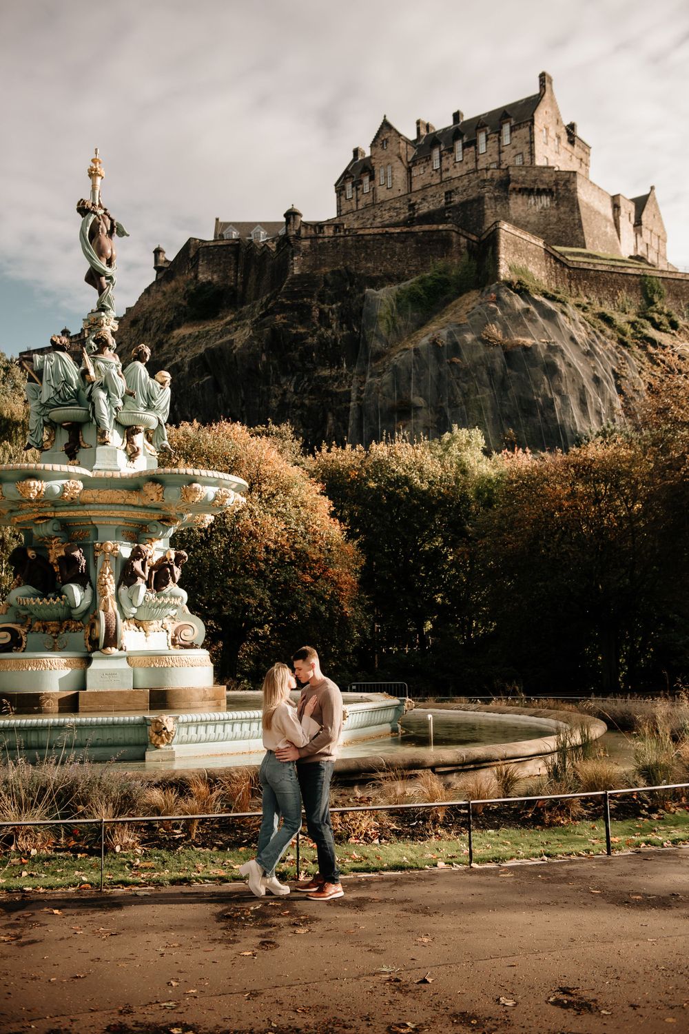 A romantic couple stands by an ornate fountain in front of the majestic Edinburgh Castle on a moody autumn day.